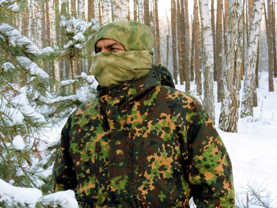 На фото человек в зимней балаклаве среди заснеженного зимнего леса.